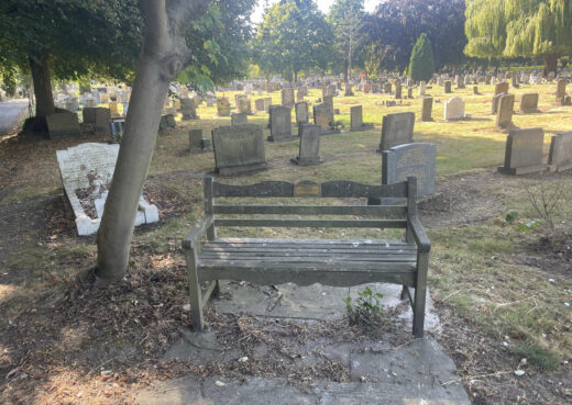 Weathered Woodcraft memorial bench among gravestones and mature trees at Chanterlands Crematorium and Northern Cemetery in Hull.