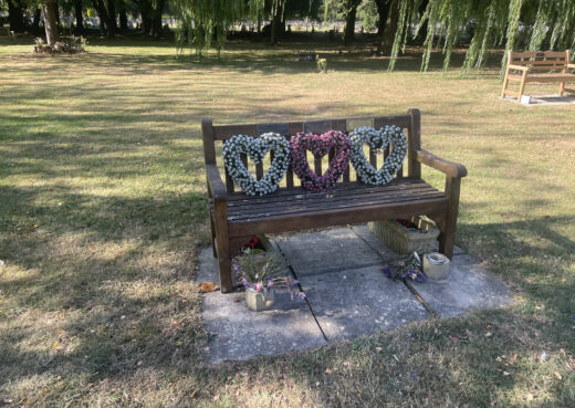 Woodcraft memorial bench decorated with three floral heart wreaths and flowers in memory of loved ones.