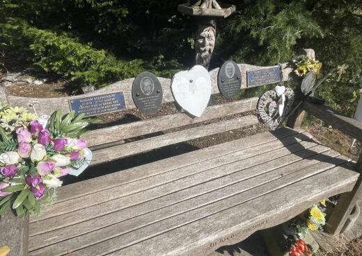 Close-up of a naturally weathered memorial bench decorated with family plaques, hearts and colourful flowers.