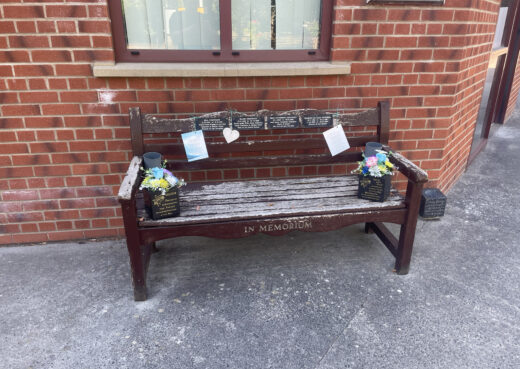 Old painted Woodcraft memorial bench decorated with flowers, plaques and remembrance notes outside Chanterlands Crematorium.