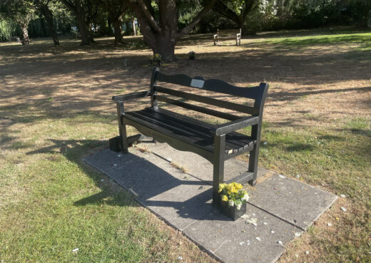 Dark-stained wooden memorial bench with flowers beneath mature trees at Chanterlands Crematorium in Hull.