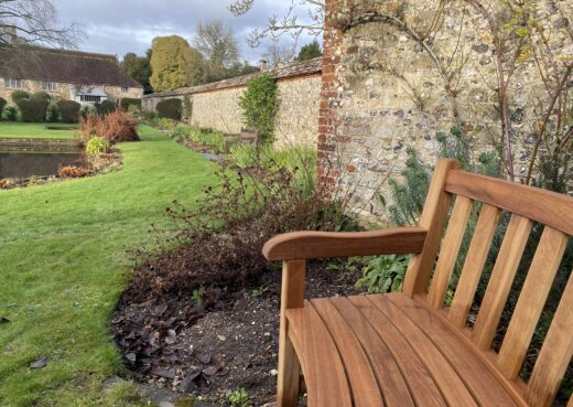 Side view of a handcrafted curved York bench showing shaped armrest and slatted seat, set within the historic gardens of the Hospital of St Cross