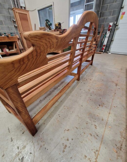 Low angle rear view of a traditional Lutyens-style hardwood bench finished in teak oil inside the Woodcraft workshop.