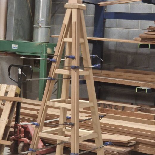 Partially assembled wooden garden obelisk held with clamps in a workshop setting.