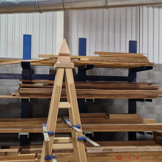 Wooden garden obelisk under construction with clamps attached, surrounded by stacked timber.