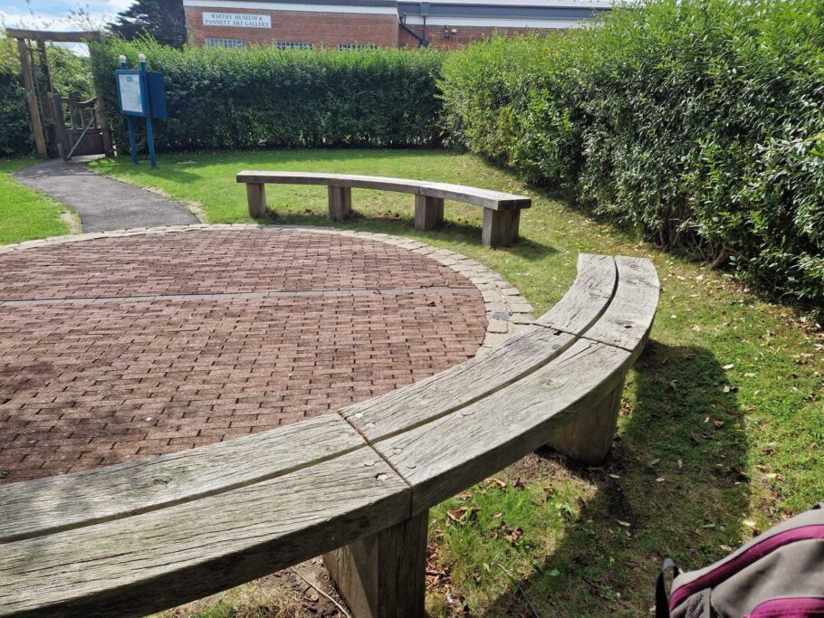Weathered curved wooden bench at Pannett Park with Whitby Museum and Pannett Art Gallery in the background