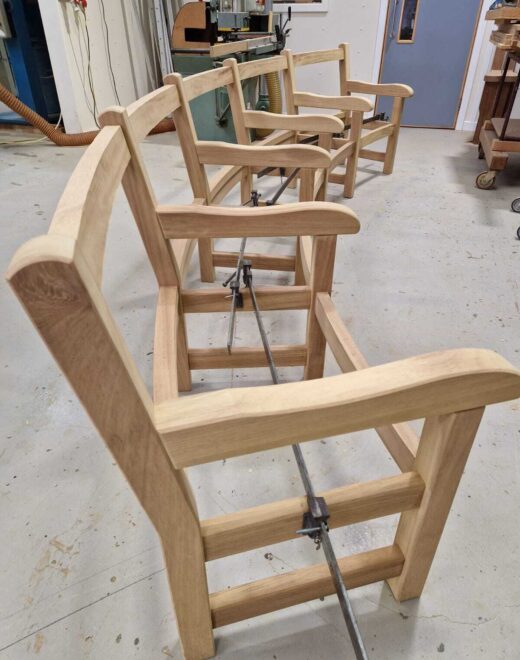 Curved hardwood bench frames from side view in the Woodcraft UK workshop during construction, highlighting the flowing back rails and handcrafted arm details before final assembly